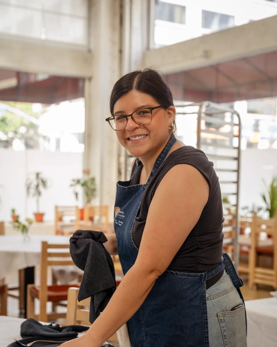 Server in a denim apron smiling while resetting a table in a bright dining room